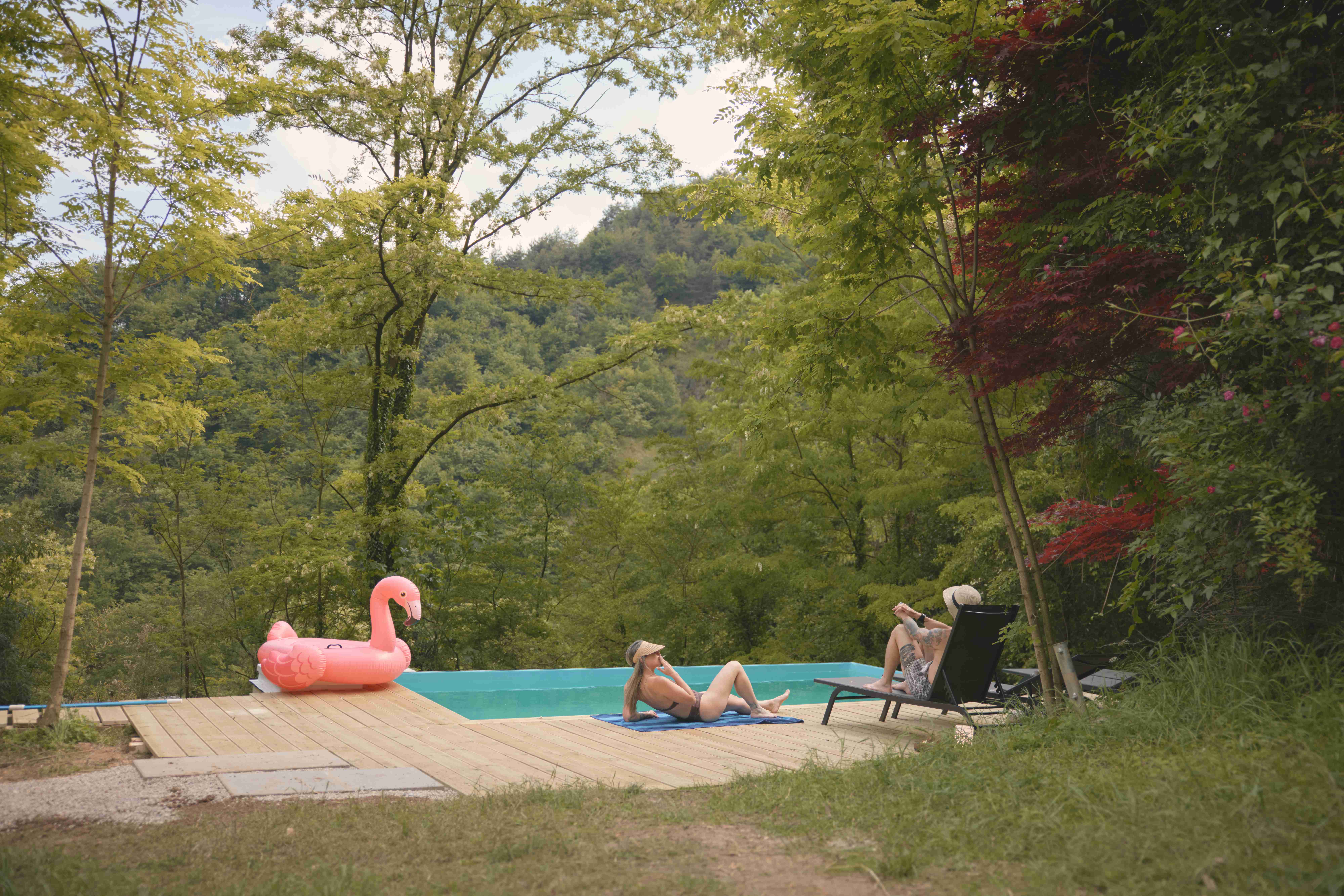 Wooden pool deck surrounded by lush forest with a flamingo float and lounge chairs